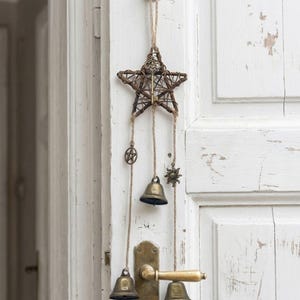 Puede incluir: Un adorno de pared rústico en forma de estrella hecho de ramitas tejidas, adornado con pequeñas campanas y amuletos de latón. La estrella está suspendida por un cordón de hilo natural, con tres campanas colgando debajo. La puerta es blanca con pintura desgastada.