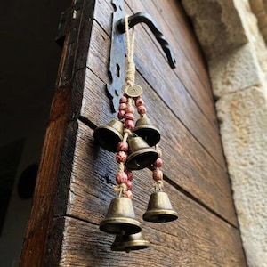 Peut inclure: Une cloche décorative suspendue à une porte en bois. La cloche est composée de petites cloches de couleur bronze, enfilées avec des perles rouges et de la ficelle. Une poignée de porte en métal et une plaque décorative sont visibles.