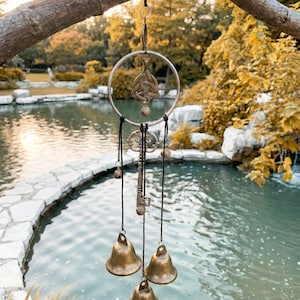 May include: A hanging wind chime with a Celtic knot design, a key, and three bronze bells. The wind chime is suspended from a tree branch with a black ribbon. The background features a pond and trees.
