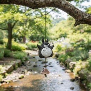 Puede incluir: Un carillón caprichoso con un personaje de dibujos animados en blanco y negro y un badajo en forma de hongo. El carillón está suspendido de una rama de árbol contra un fondo borroso de un arroyo y exuberante vegetación.