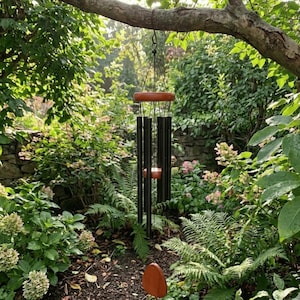 Puede incluir: Un carillón de viento negro con una parte superior y detalles de madera cuelga de una rama de árbol en un jardín. El carillón tiene varios tubos largos y oscuros y una pequeña pieza redonda de madera en el medio. Una pequeña pieza marrón en forma de lágrima cuelga en la parte inferior.