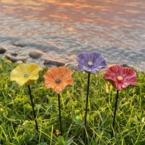 May include: Four ceramic flower garden stakes in yellow, orange, purple, and red. Each flower has a detailed centre. The stakes are set in green grass with water in the background. These decorative flowers are a garden accent, perfect for adding colour.