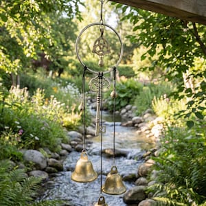 以下が含まれることがあります： 金属製のリング、ケルトノット、キー、真鍮製のベル3つを備えた装飾的なウィンドチャイム。ウィンドチャイムは黒いコードから吊り下げられており、小川と緑豊かな緑のぼやけた背景が特徴です。