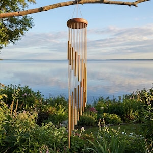 May include: A decorative wind chime with a wooden top and numerous golden tubes hanging from strings. The chime is suspended from a tree branch, set against a backdrop of a lake and lush greenery. The tubes are arranged in a spiral design.