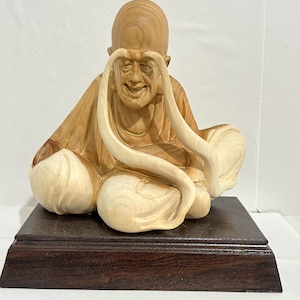 Peut inclure: Une sculpture en bois clair représentant un personnage chauve aux longs sourcils incurvés tombant vers le bas, assis en tailleur. Le personnage porte une robe et est fixé sur un socle rectangulaire en bois brun foncé à la finition lisse.
