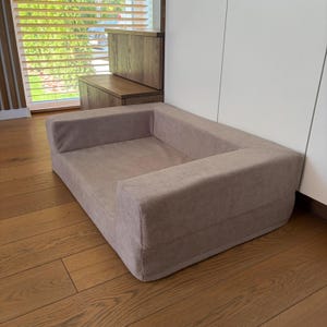 May include: A rectangular pet bed with a soft, light brown, suede-like fabric finish. The bed features a low-profile design with raised, padded side bolsters that form a supportive border around the central sleeping area, creating a cozy enclosure.