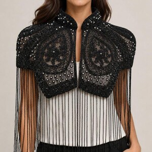 Peut inclure: Un boléro noir orné de perles et de broderies complexes sur une base en maille transparente. Le vêtement présente un col montant, des épaules structurées et de longues franges de perles noires délicates suspendues à l'ourlet.