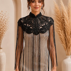 May include: A black beaded capelet featuring a high collar and intricate floral-inspired embroidery. The garment is adorned with long, thin black fringe that extends from the shoulders and chest, creating a layered, textured appearance.