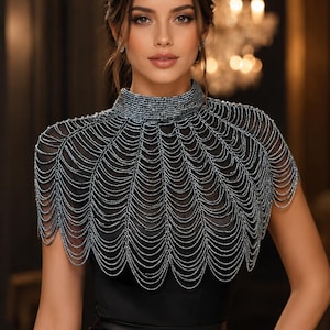 May include: A high-neck beaded capelet featuring multiple layers of draped, shimmering dark grey chains that form a scalloped pattern over the shoulders and chest, worn over a solid black sleeveless garment with a smooth, satin-like finish.