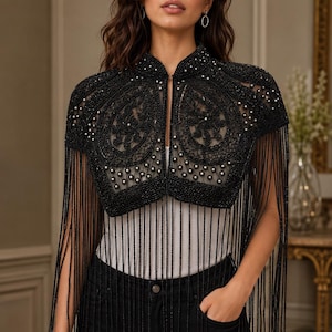 May include: A black beaded bolero jacket featuring intricate embroidery, a high collar, and long fringe detailing. The garment is adorned with small white pearls and black beads arranged in a paisley-inspired pattern over a sheer mesh base.
