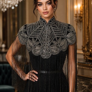 Könnte beinhalten: Ein schwarzes Kleid mit hohem Kragen und einem aufwendigen, goldfarbenen Perlenbesatz über Schultern und Brust. Das Kleidungsstück verfügt über lange, feine schwarze Fransen, die von der bestickten Passe herabhängen und einen Lagen-Look erzeugen.