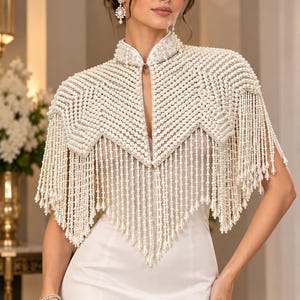 May include: A white pearl-embellished capelet featuring a high collar and intricate geometric patterns of small round pearls. The garment is adorned with long, cascading fringe made of pearl strands ending in teardrop-shaped pearl accents.