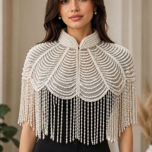May include: A white pearl-embellished capelet featuring a high collar and scalloped edges. The garment is adorned with multiple rows of draped white pearls that form a layered pattern, extending into long, dangling pearl fringe strands.