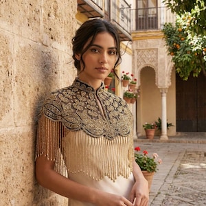 May include: A woman wearing a cream-colored dress with a high-neck, beaded cape. The cape is black and gold, with intricate floral patterns and beaded fringe. The background features a stone wall and a building with architectural details.