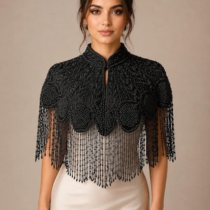 Könnte beinhalten: Ein schwarzes, perlenbesetztes Capelet mit Stehkragen und aufwendiger Blumenstickerei. Das Kleidungsstück ist mit dichten schwarzen Perlen verziert und hat einen geschwungenen Saum mit langen, herabhängenden Perlenfransen über einem hellen Kleid.