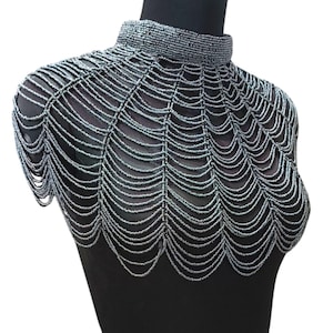 May include: A beaded collar necklace in silver, displayed on a black mannequin. The necklace features a high, rounded collar and a web-like design that drapes over the shoulders. The beads are small and evenly spaced, creating a delicate, yet structured look.