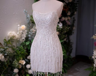 May include: A strapless white mini dress featuring an intricate pattern of silver sequins and clear beads. The bodice is fitted, and the skirt is adorned with long, dangling beaded fringe that hangs from the hemline, creating a textured effect.