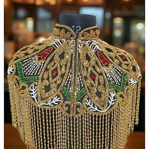 May include: Black beaded collar with gold fringe. The collar features intricate gold, red, green, and white beadwork in a floral and geometric pattern. The collar is displayed on a black mannequin head.