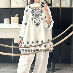 May include: A white tunic featuring black geometric embroidery around the neckline and hem, paired with matching white wide-leg trousers that have horizontal black striped detailing at the cuffs and a coordinating white scarf draped over the shoulders.