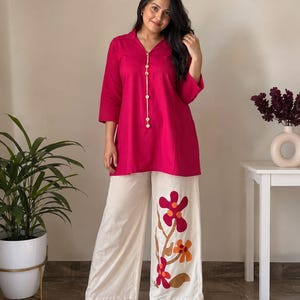 May include: A bright fuchsia tunic with a collared neckline and a vertical row of light-coloured buttons, paired with wide-leg off-white trousers featuring a large, colourful floral applique design in shades of red, orange, and brown on one leg.