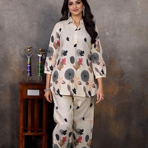May include: Cream-colored floral print pajama set. The top has a collared neckline and three-quarter sleeves. The pants are wide-legged. The floral pattern includes black, pink, and brown elements. The set is made of a lightweight fabric.