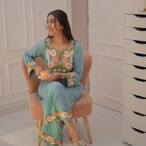 May include: A light blue embroidered two-piece outfit. The top has floral embroidery around the neckline and sleeves. The trousers have matching floral embroidery along the hem. The outfit is paired with patterned sandals.
