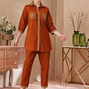 Op de afbeelding: Roestkleurige tweedelige outfit met een kraag, shirt met lange mouwen met geborduurde details op de zakken en het midden. De bijpassende broek heeft een rechte pijp. Transparante hakken maken de look compleet.