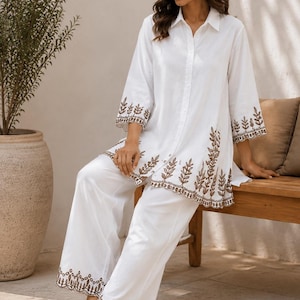 Könnte beinhalten: Weißes zweiteiliges Outfit mit einem Hemd mit Knöpfen und einer Hose mit weitem Bein. Das Hemd und die Hose sind mit braunen, gestickten Blattmustern an den Rändern verziert. Das Outfit wird mit beigen Sandalen kombiniert.