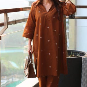 May include: A woman wearing a rust-colored, two-piece outfit with a tunic top and matching pants. The tunic has a V-neck and diamond-shaped white embroidery. She carries a brown handbag and wears sunglasses and sandals. The outfit is suitable for casual wear.