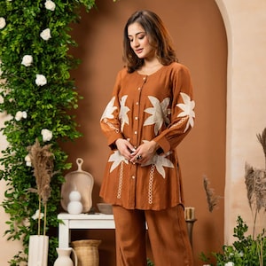 May include: A rust-colored long-sleeve button-down tunic and matching wide-leg trousers. The tunic features white embroidered palm leaf motifs on the sleeves and front, a round neckline, and a full-length front button closure with white buttons.