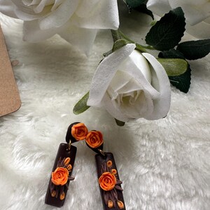 Puede incluir: Pendientes hechos a mano con una base rectangular marrón oscuro, adornados con detalles de rosas y hojas naranjas. Los pendientes tienen ganchos dorados y están colocados sobre un fondo blanco y esponjoso con rosas blancas.
