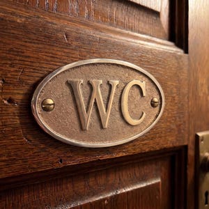 Könnte beinhalten: Nahaufnahme einer Holztür mit einem ovalen Messingschild mit der Aufschrift "WC". Das Schild ist mit zwei kleinen Schrauben befestigt. Ein Messing-Türknauf und Beschläge sind ebenfalls sichtbar.