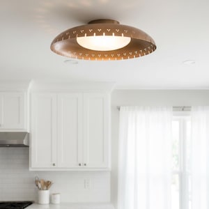 May include: A bronze-colored, mid-century modern flush mount ceiling light fixture. The light has a perforated shade with a white, frosted glass diffuser. The fixture is mounted on a white ceiling in a kitchen setting.