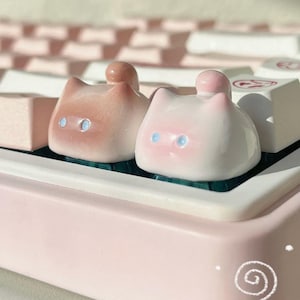 Puede incluir: Primer plano de dos teclas con forma de gato en tonos rosa y blanco, con ojos azules y una pequeña bola en la parte superior. Las teclas están sobre un teclado rosa con teclas blancas y rosas. El diseño es lindo y caprichoso.