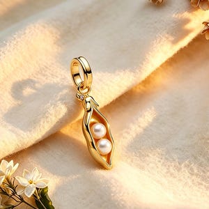 Könnte beinhalten: Ein goldener Anhänger in Form einer Erbsenschote, die zwei weiße, kugelförmige Perlen enthält. Der Anhänger verfügt über eine kleine, kreisförmige Öse an der Oberseite zur Befestigung an einer Kette und eine polierte Oberfläche, die das Licht reflektiert.