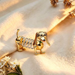 May include: A gold-toned dachshund figurine featuring a metallic spring body connecting the front and back sections. The dog has long ears, black eyes, a black nose, and a small green collar detail. The figure stands on four small legs.