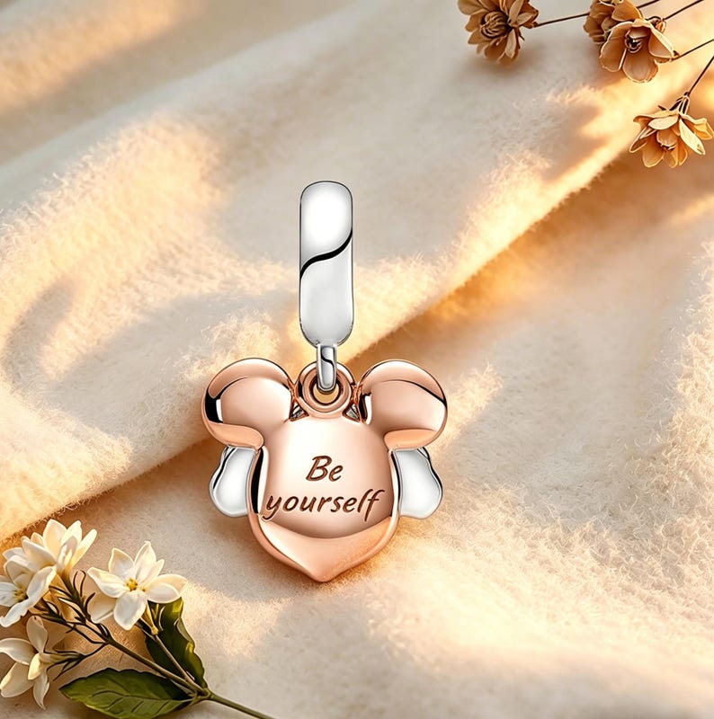 Op de afbeelding: Een ros&eacute; gouden en zilveren bedel met een Mickey Mouse-hoofdontwerp. De bedel heeft de tekst "Be yourself" gegraveerd op de voorkant. De bedel hangt aan een zilveren beugel.