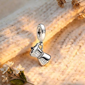 May include: A silver charm in the shape of a coffee maker, embellished with clear crystals. The charm has a loop at the top for attachment and is set against a soft, textured backdrop. The charm is a jewelry item.