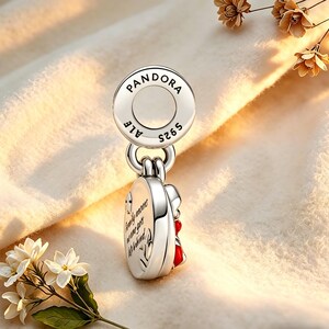 May include: A silver Pandora charm with the brand name and "S925 ALE" markings. The charm features a heart-shaped design with red accents and the inscription "Family moments are our greatest gifts left behind."