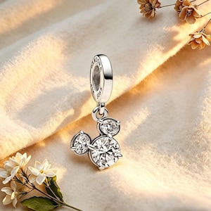 May include: A silver charm with a Mickey Mouse design. The charm features three clear, sparkling cubic zirconia stones arranged in the shape of Mickey Mouse's head. The charm is suspended from a silver loop with the word "Pandora" engraved on it.