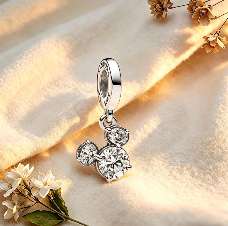 May include: A silver charm with a Mickey Mouse design. The charm features three clear, sparkling cubic zirconia stones arranged in the shape of Mickey Mouse's head. The charm is suspended from a silver loop with the word "Pandora" engraved on it.