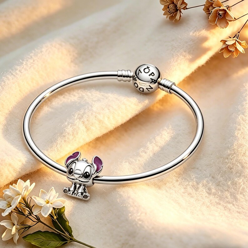 May include: A silver bracelet with a charm featuring the character Stitch from Lilo & Stitch. The charm is detailed with purple accents on the ears. The bracelet is displayed on a soft, cream-colored surface.
