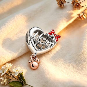 May include: A silver charm with the word "Mom" in a heart-shaped design, accented with a red bow and a dangling charm. The charm has a rose gold-colored Minnie Mouse-shaped charm hanging from it. The charm is on a soft, light-colored surface.