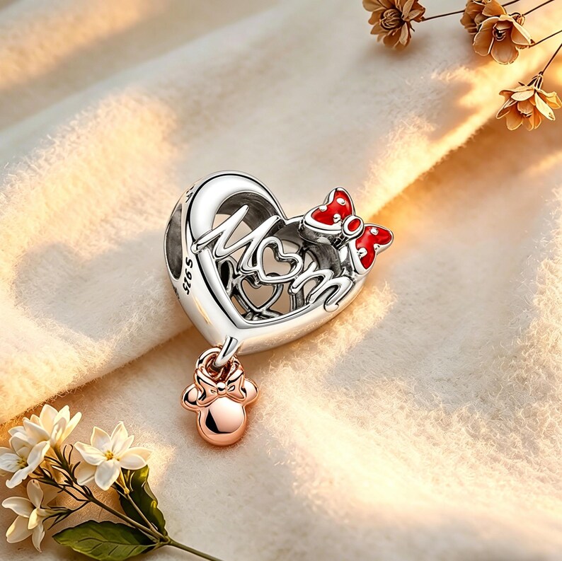 May include: A silver charm with the word "Mom" in a heart-shaped design, accented with a red bow and a dangling charm. The charm has a rose gold-colored Minnie Mouse-shaped charm hanging from it. The charm is on a soft, light-colored surface.