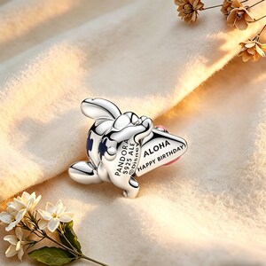 May include: A silver charm depicting a cartoon character with blue details and the words "Aloha Happy Birthday." The charm is from the Pandora Disney collection.