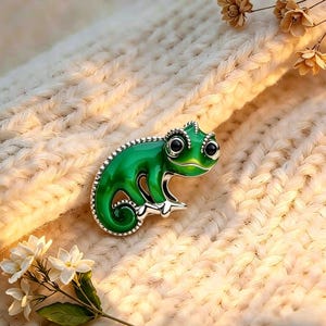 May include: A green chameleon brooch with a silver border and black eyes. The brooch is set against a cream-coloured, textured background with small white flowers and dried floral accents.