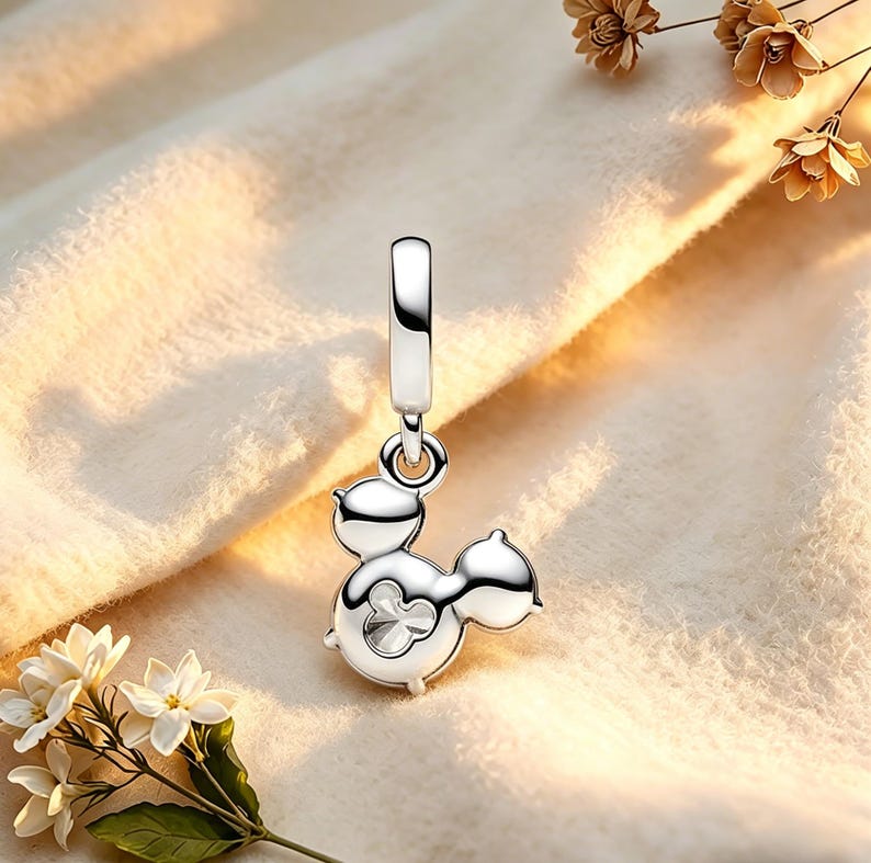 May include: A silver charm pendant in the shape of a cartoon character with a Mickey Mouse-like design. The charm has a loop at the top for hanging and a small, clear gem in the center. The charm is displayed on a soft, cream-coloured fabric.