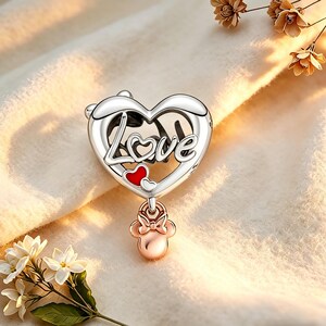 May include: A silver charm with a heart-shaped design. The word "Love" is written across the heart. Two small red hearts are inside the heart. A rose gold charm in the shape of a bow and Minnie Mouse's head hangs below the heart.