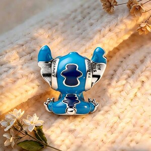 May include: A silver charm shaped like the character Stitch, featuring bright blue enamel detailing on the ears and body, with dark blue accents. The side of the ear is engraved with the text Disney, Pandora, and S925 ALE, indicating metal purity.