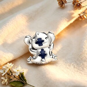 May include: A silver charm depicting a cartoon character with large ears and a blue chest detail. The charm has small feet and hands, and the word "PANDORA" is engraved on the base. The charm is set against a soft, neutral background.